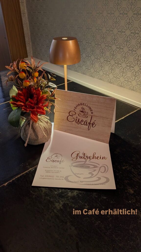 Gutschein des Fehrbelliner Eiscafés auf einem Tisch, dekoriert mit moderner Lampe und herbstlichem Blumenarrangement.